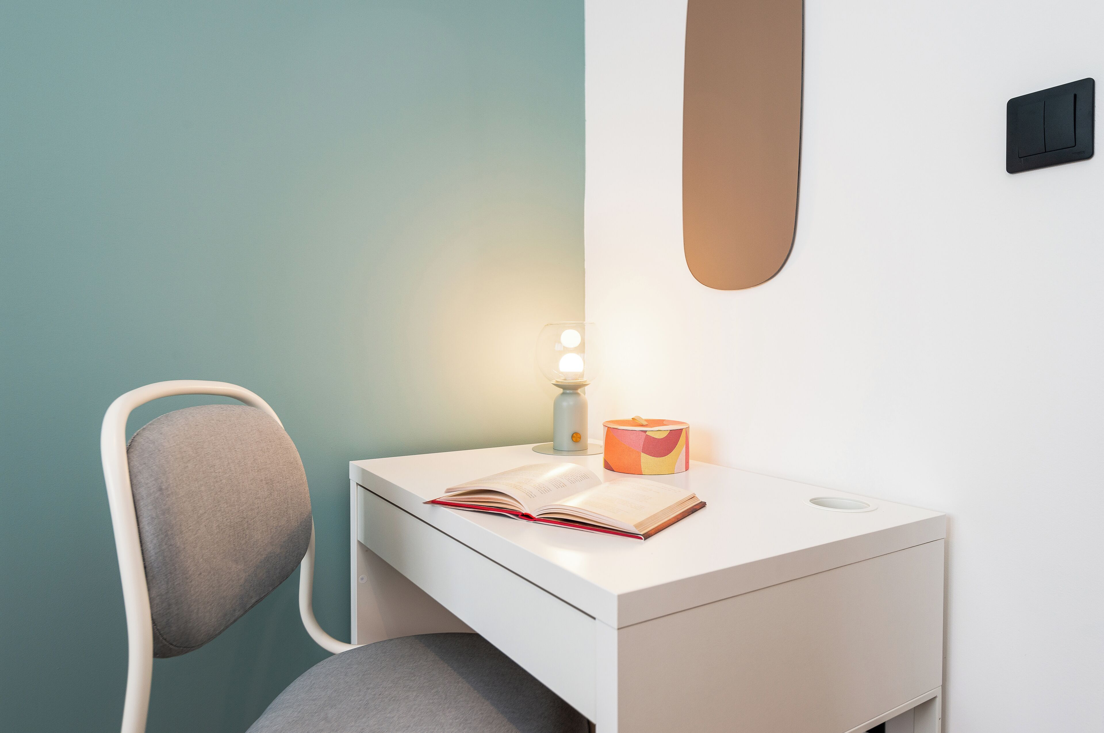 Un espacio de trabajo pequeño y moderno con un escritorio blanco, una silla gris, un libro, una lámpara y una caja de colores.