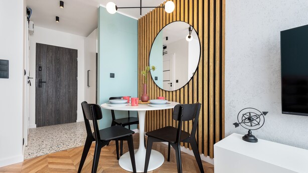 A modern dining area with a round table, two black chairs, a wooden slat wall, a round mirror, and a television on a white stand. - Szafir - by Kairos Apartments (Gliwice)