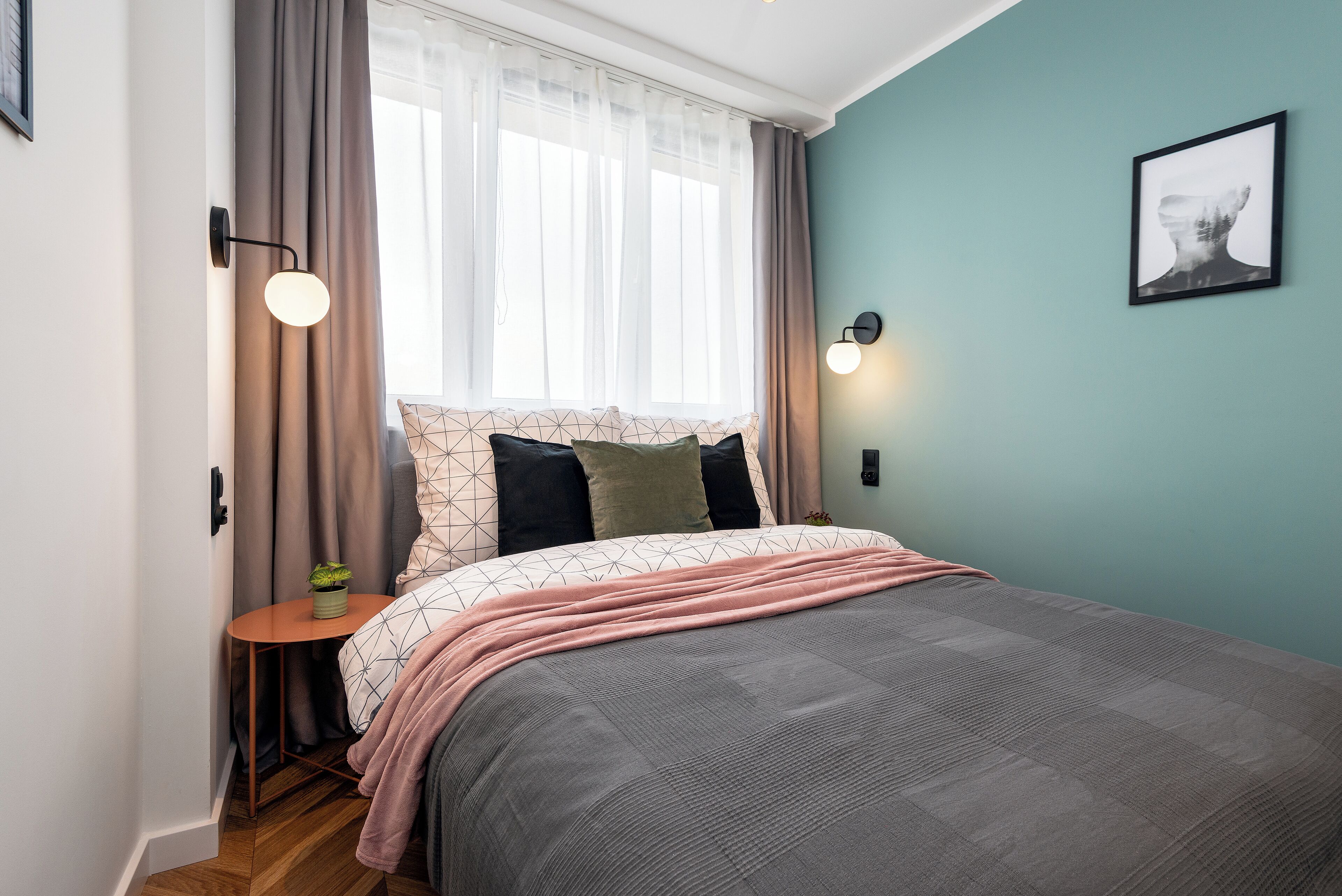 A bedroom with a bed, a bedside table with a lamp, a window with curtains, and a wall-mounted light fixture.