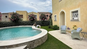 Zona de piscina con una piscina circular, escalones que bajan al agua y dos sillones de mimbre con una mesa pequeña y girasoles.