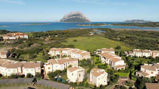 Una zona residencial costera con casas escalonadas, una colina rocosa destacada al fondo y una vista despejada del mar.