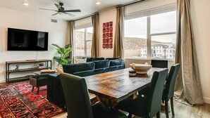 A modern living room with a wooden dining table, dark sofas, a flat-screen TV, and a large window with curtains.