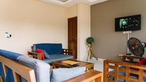 A living room with a sofa, a wooden coffee table, a TV mounted on the wall, and a standing fan.