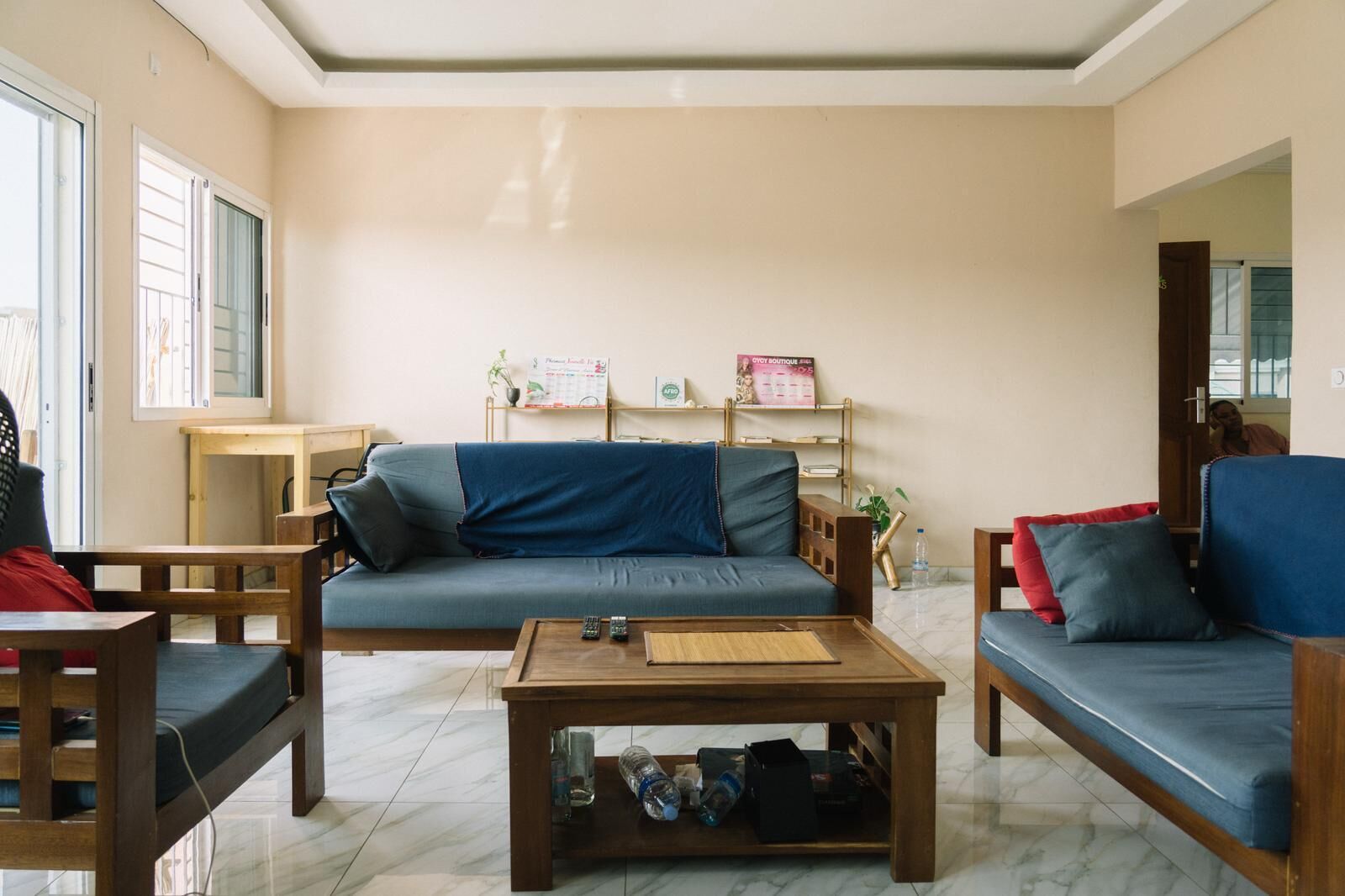 Una sala de estar con un sofá, dos sillones, una mesa de centro y una pequeña mesa de madera. Hay un calendario en la pared y una puerta que da al exterior.