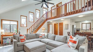 A spacious living room with a sectional sofa, ottoman, and a wooden staircase leading to an upper level. - 3 Mi to Tobyhanna State Park: Home w/ Serene Deck! (Tobyhanna)