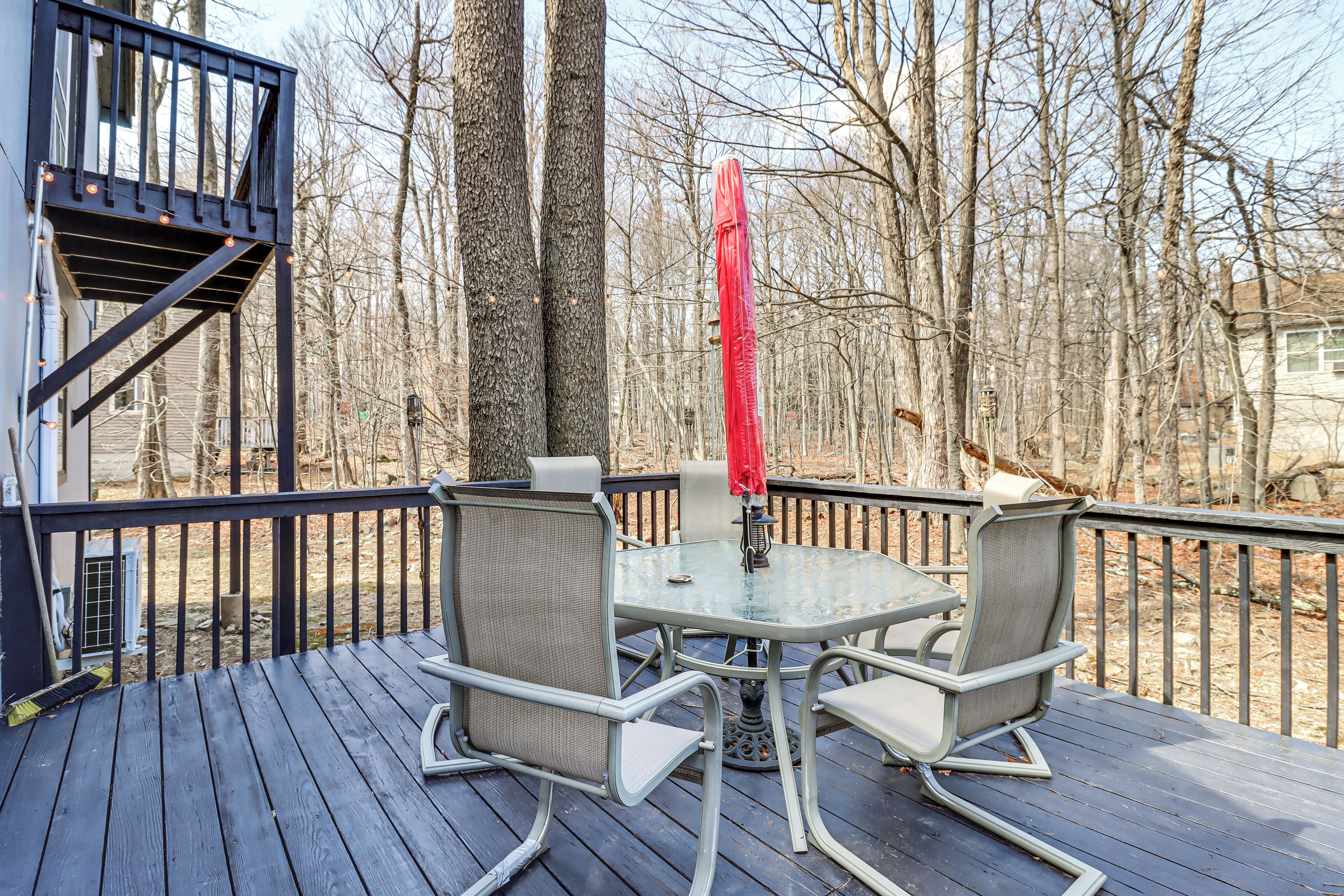 A deck with a glass table and chairs, a red umbrella, and a view of leafless trees.