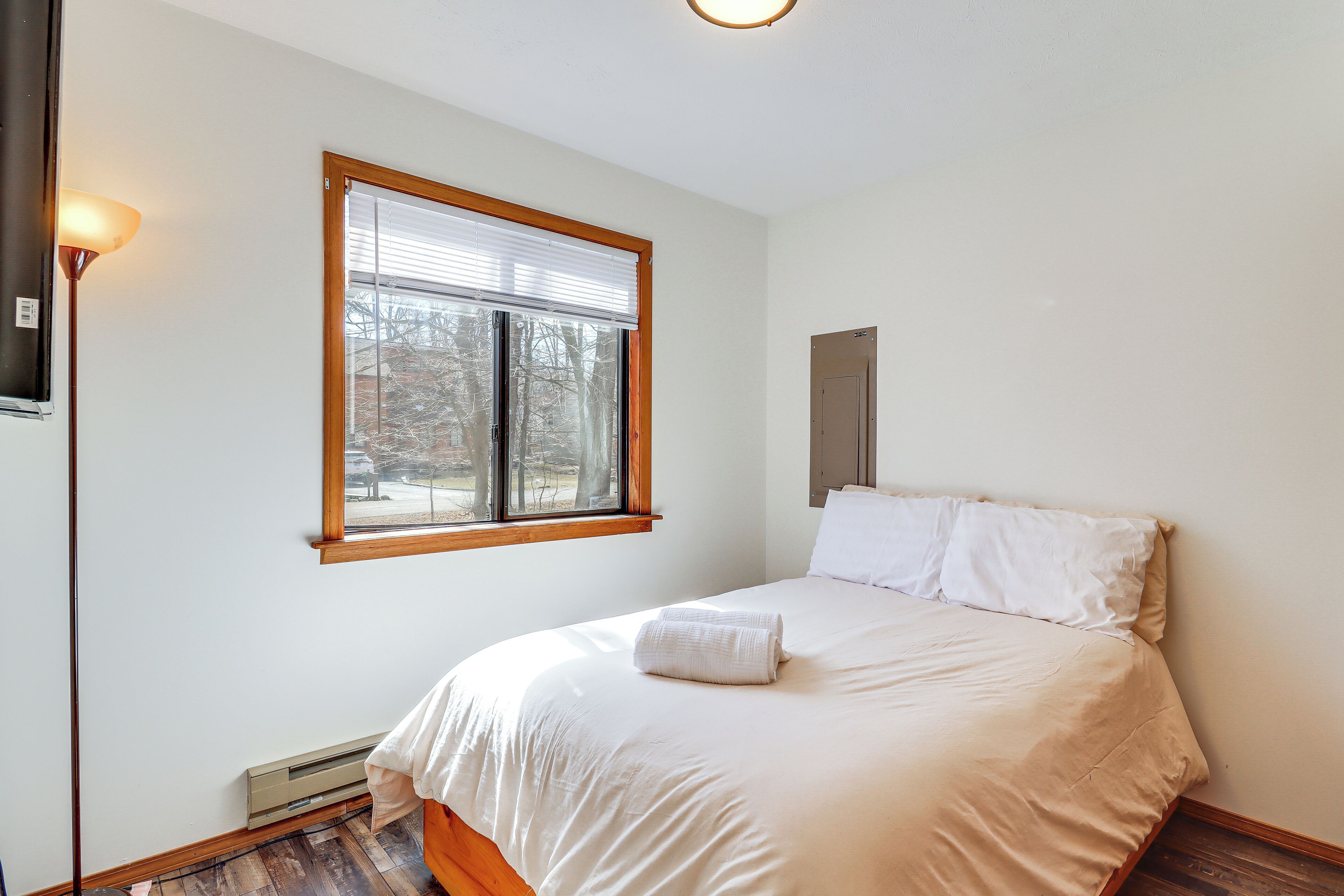 A bedroom with a bed, a window with blinds, a wall-mounted heater, and a light fixture.