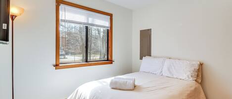 A bedroom with a bed, a window with blinds, a wall-mounted heater, and a light fixture.