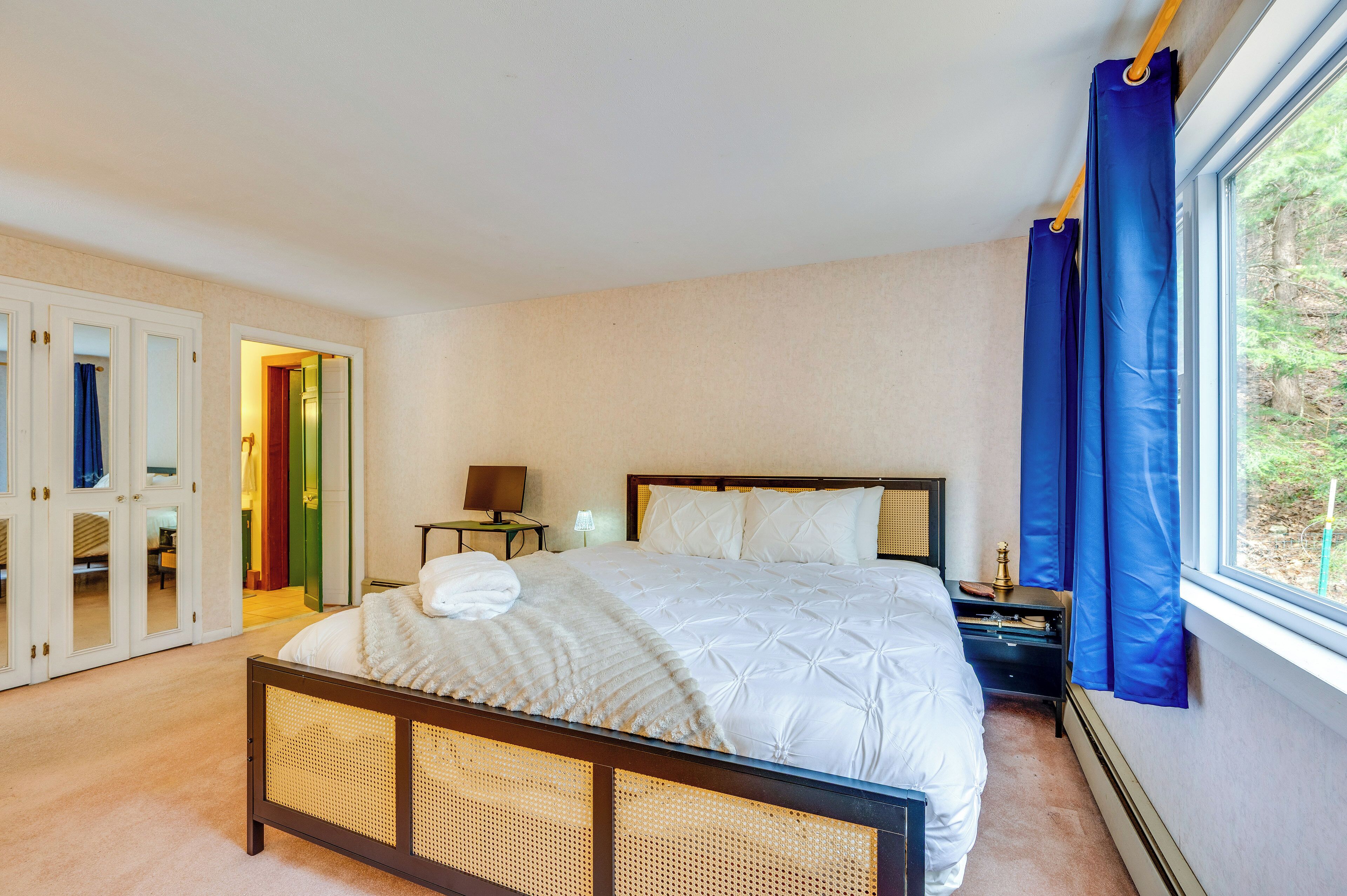 A bedroom with a bed, bedside table, lamp, and a view of greenery through a window.