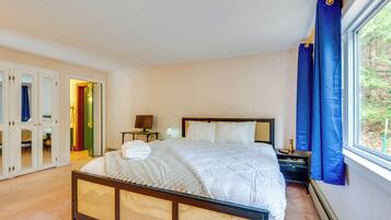 A bedroom with a bed, bedside table, lamp, and a view of greenery through a window.