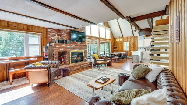 A spacious living room with a fireplace, leather sofas, wooden flooring, and large windows offering a view of the outdoors.