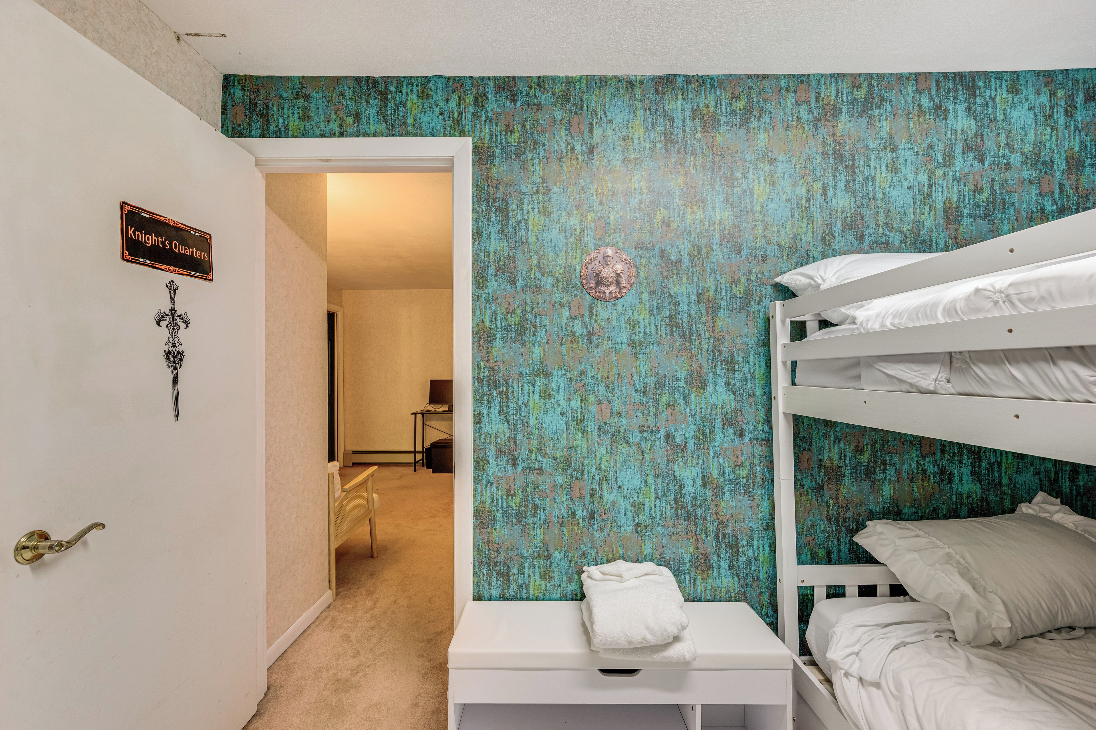 A bedroom with bunk beds, a white bedside table, a wall with a teal textured mural, and a door labeled "Knight's Quarters".