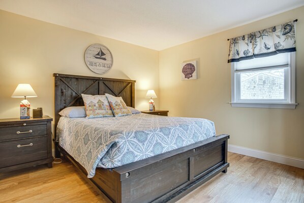 A bedroom with a wooden bed, two bedside tables with lamps, a wall clock, and a window with a view of a house.