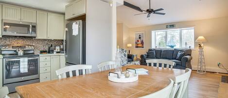 A modern kitchen with a dining area, featuring a large wooden table, white chairs, and a ceiling fan.