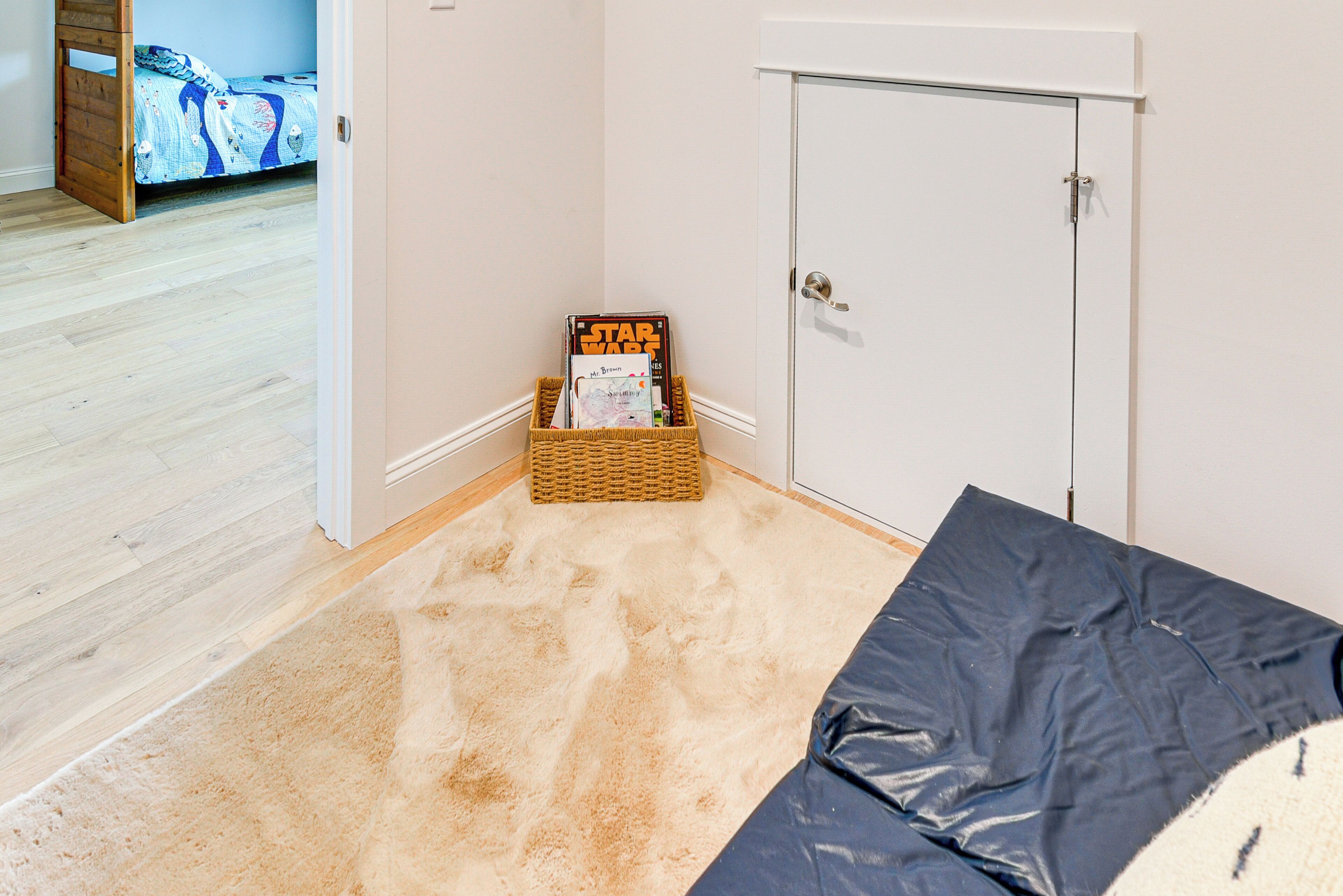 A room with a wooden floor, a white door, and a corner with a carpet and a basket containing magazines.