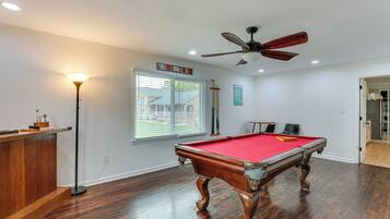 A pool table with a red felt surface, a ceiling fan, and a window with blinds.