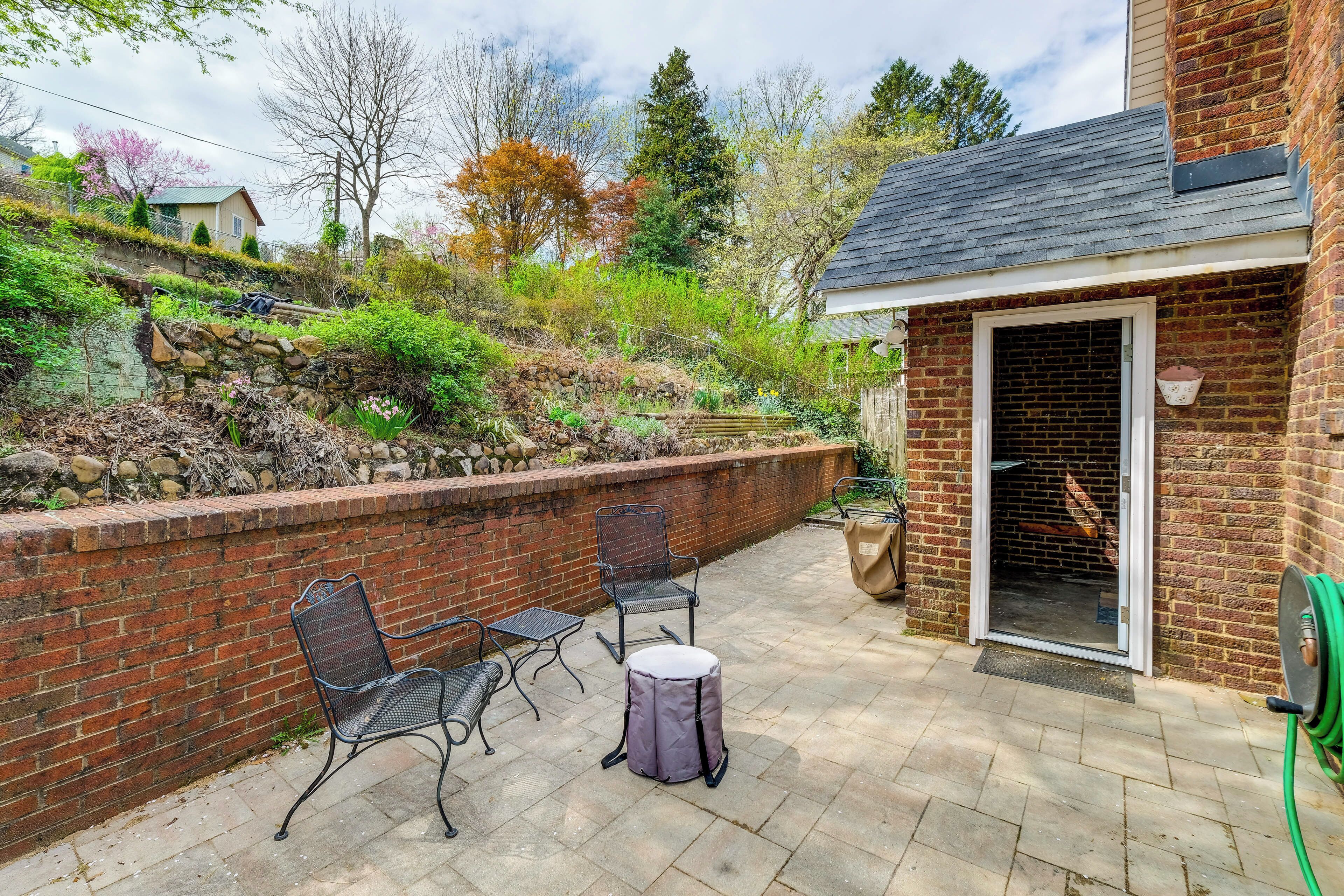 An outdoor patio with a brick wall, two metal chairs, a small round table, and a green garden hose.