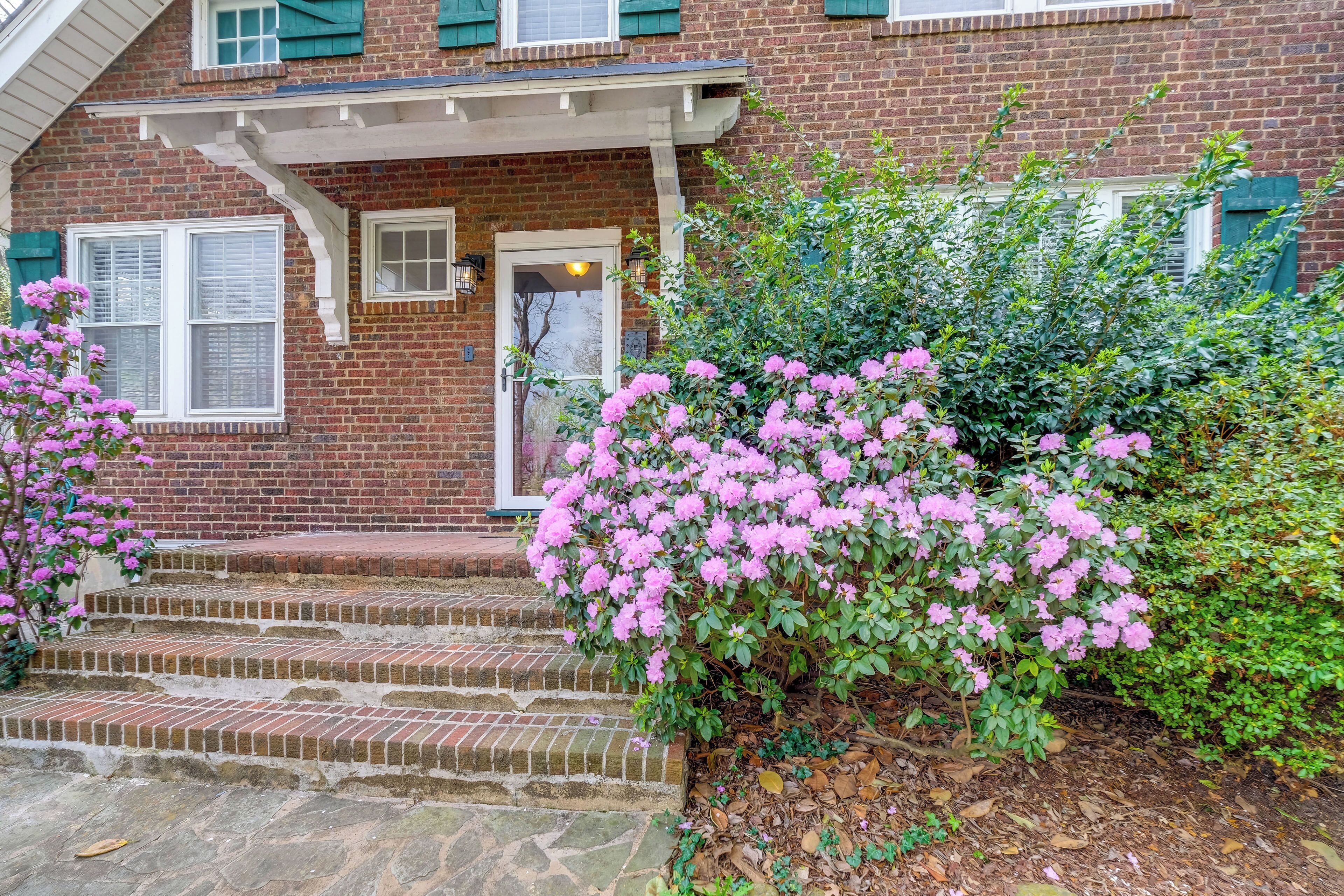 A brick house with a front porch, steps leading to the entrance, and a bush with pink flowers.