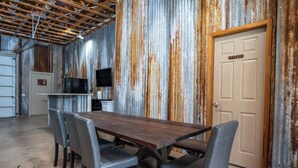A rustic dining area with a wooden table and grey upholstered chairs, a television, and a storage door. - King Beds, Game Room, On 290, Large Estate,Kitchen (Fredericksburg)