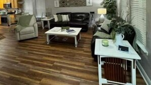 A spacious living room with hardwood flooring, a ceiling fan, a black leather sofa, a white coffee table, and a dining area with a small table and chairs. - Serenity Cottage (Ocean Springs)