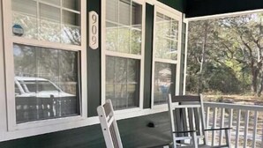 A porch with white rocking chairs and a small table, a green exterior wall, and a window with a number '9' on it. - Serenity Cottage (Ocean Springs)