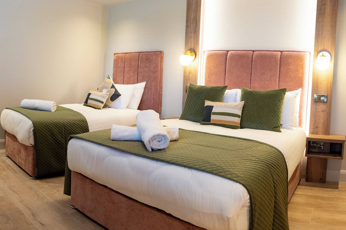 Ein Hotelzimmer mit zwei Betten, jeweils mit Kopfstütze, einem Holznachttisch und einer an der Wand montierten Lampe.