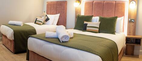 Habitación de hotel con dos camas, cada una con cabecero, mesita de noche de madera y lámpara de pared.