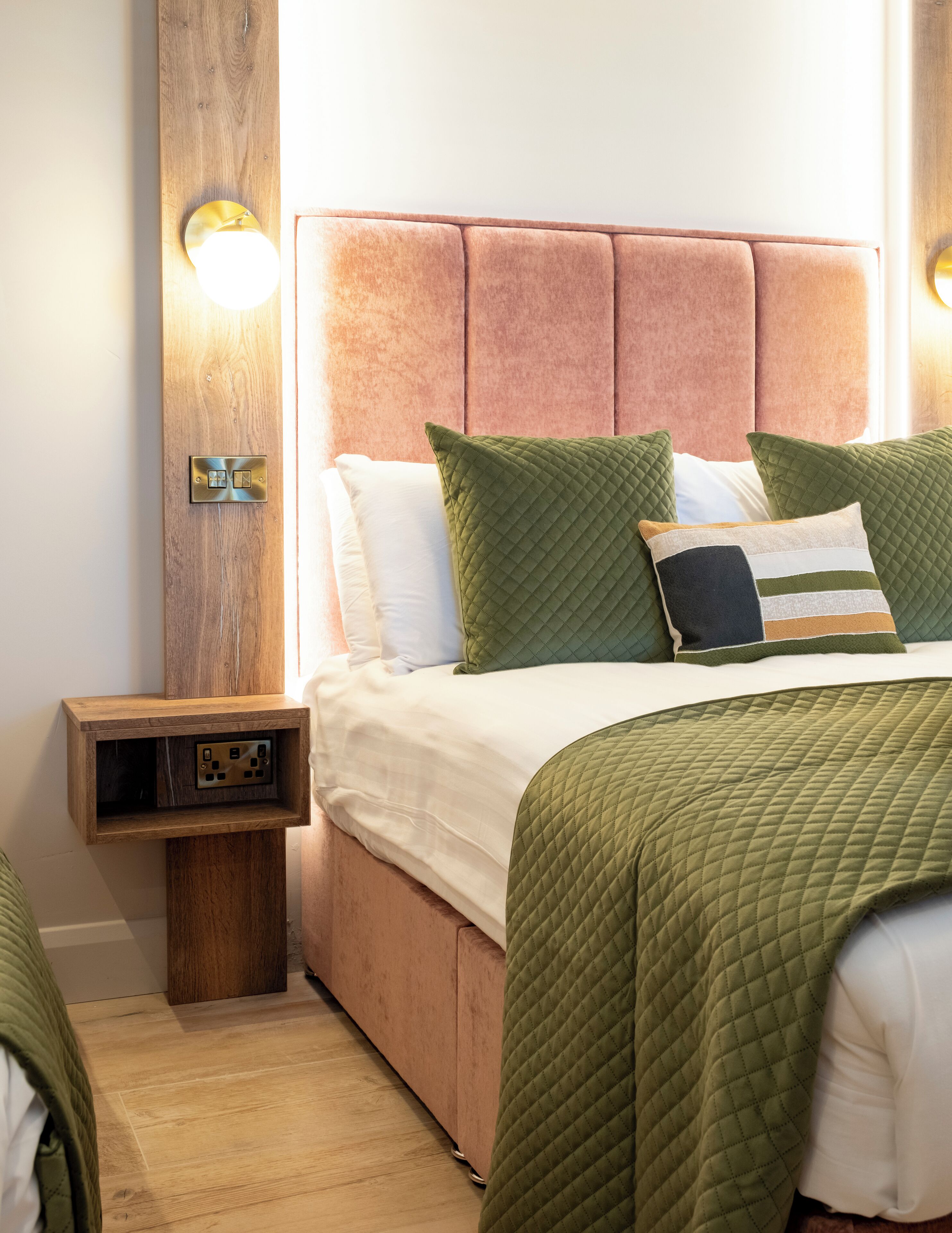 A neatly made bed with green and white bedding, a wooden nightstand with a power outlet, and a wall-mounted light fixture.