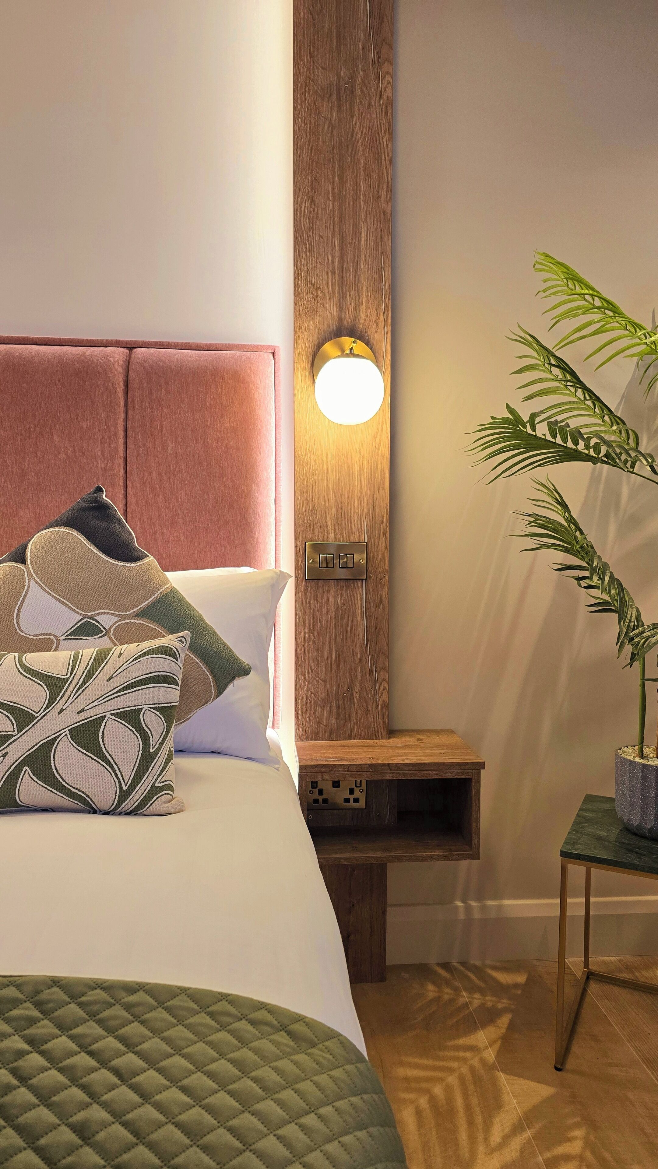 Una cama bien hecha con almohadas estampadas, una mesita de noche con una lámpara y una planta en maceta.