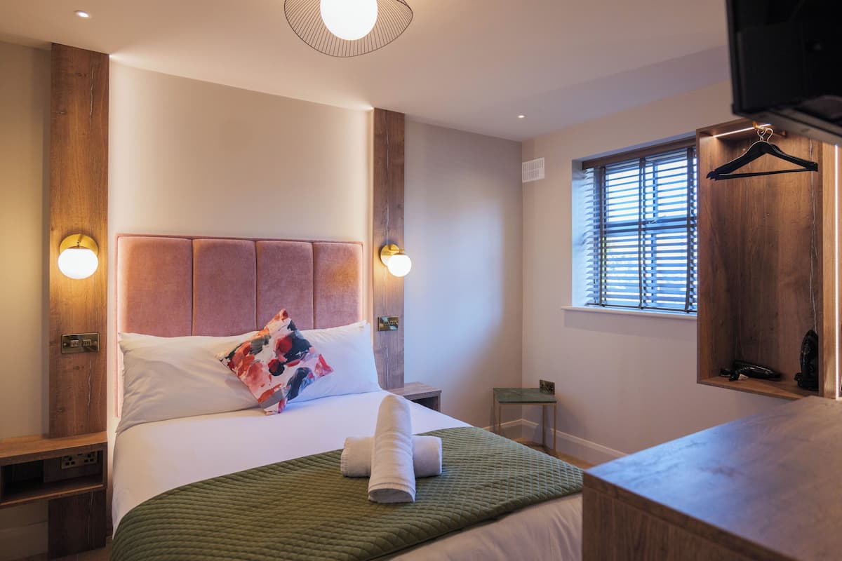 Ein ordentlich bezogenes Bett mit grüner Decke und gerolltem Handtuch, ein Holz-Kopfteil und zwei an der Wand befestigte Lampen.