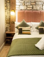 Una cama bien hecha con ropa de cama verde, una mesita de noche de madera y una pared de piedra con un panel de madera encima.