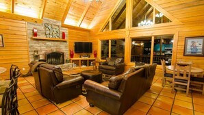 A log cabin interior with a fireplace, leather sofas, a dining area, and a kitchen visible through large windows. - Talako Lodge|4+BD|4BTH|16 Guest|Hot Tub|3 Story|Pool table (Broken Bow)