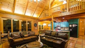 A spacious living area with leather sofas and a central ottoman, wooden ceiling beams, and a kitchen with teal cabinets. - Talako Lodge|4+BD|4BTH|16 Guest|Hot Tub|3 Story|Pool table (Broken Bow)