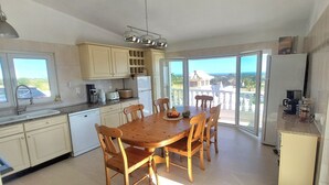 A kitchen with a dining table, chairs, cabinets, and a view through large windows. - Casa da Amizade - Tavira (Horta)