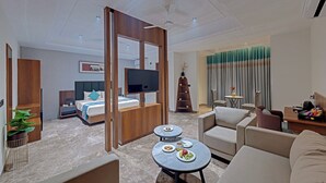A modern hotel room with a bed, a TV, a dining table with plates, and a seating area with a sofa and armchairs.