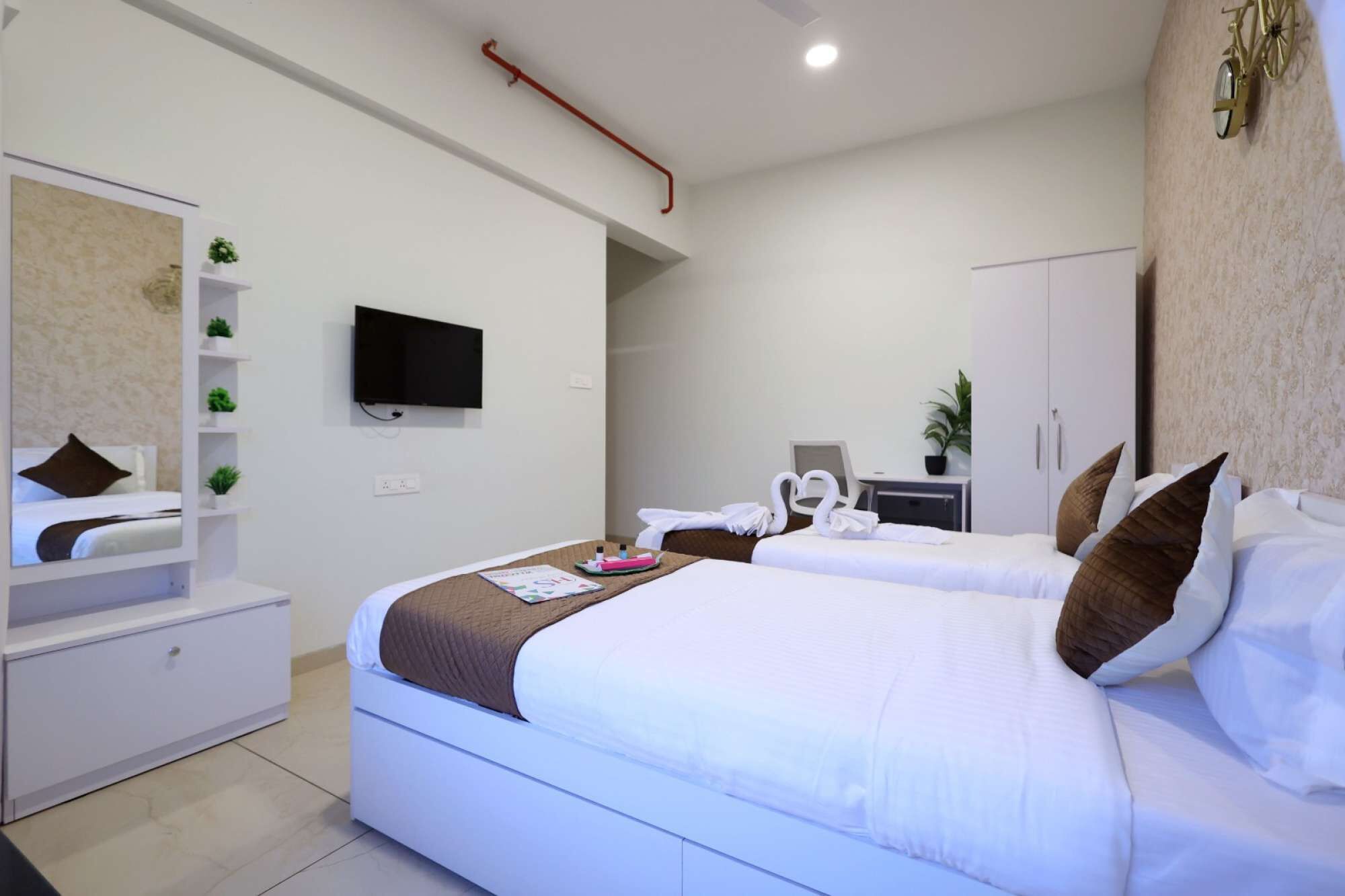 A hotel room with two beds, a TV mounted on the wall, a desk with a chair, and a wardrobe.