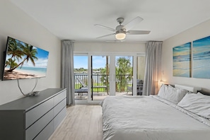 Un dormitorio con cama, televisor, ventilador de techo y balcón con vistas a palmeras y una playa.
