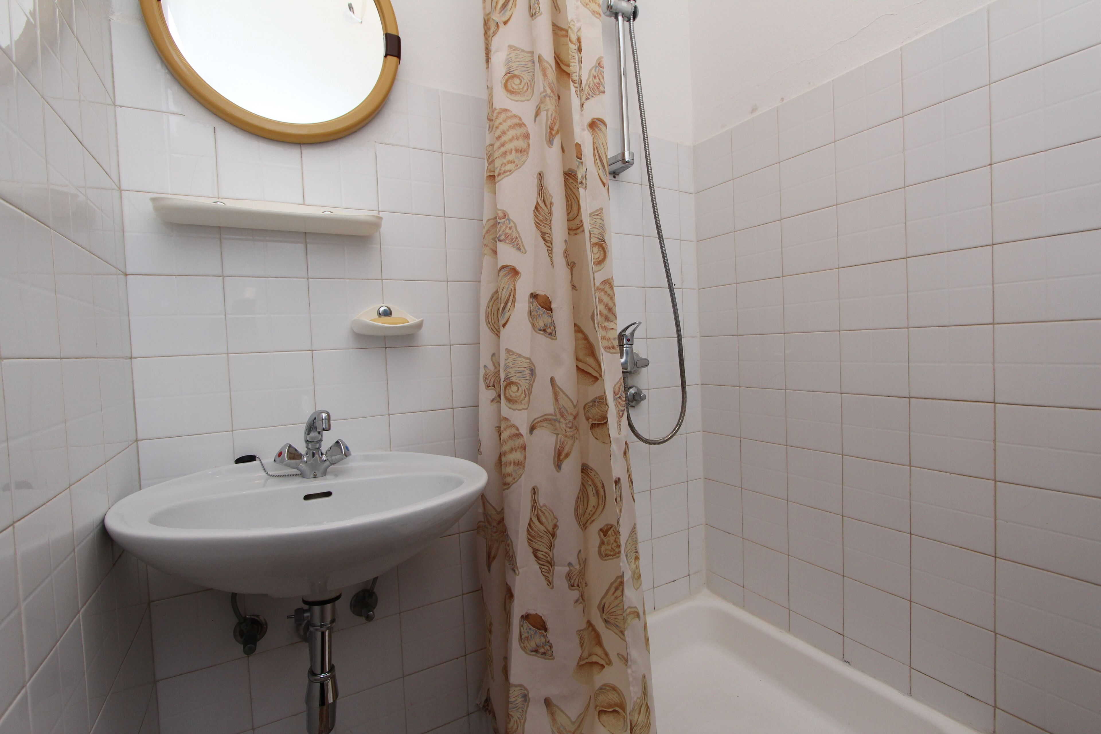 Een badkamer met een wit wastafelmeubel, een spiegel, een douche met een decoratieve gordijn en een betegelde muur.
