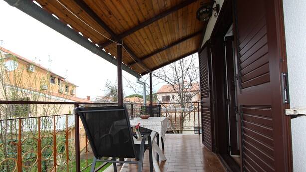 Un balcone coperto con tavolo e sedie, vista su edifici e una porta con persiana metallica.
