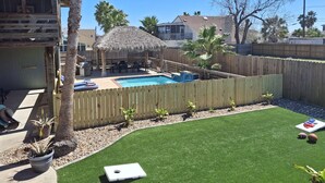 Ein Garten mit Swimmingpool, einer Holzterrasse, einem Gazebo mit Strohdach und einem eingezäunten Bereich mit Kunstgrashalm und Topfpflanzen.