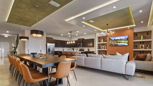 Een moderne keuken met donkere kastjes, een eethoek met een lange tafel en oranje stoelen, en een woonruimte met een bank en een televisie aan de muur.