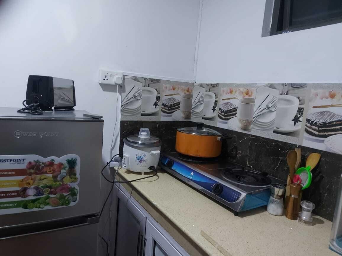 Una cocina con estufa, cocedor de arroz y microondas. Hay varios utensilios de cocina y un mueble pequeño.