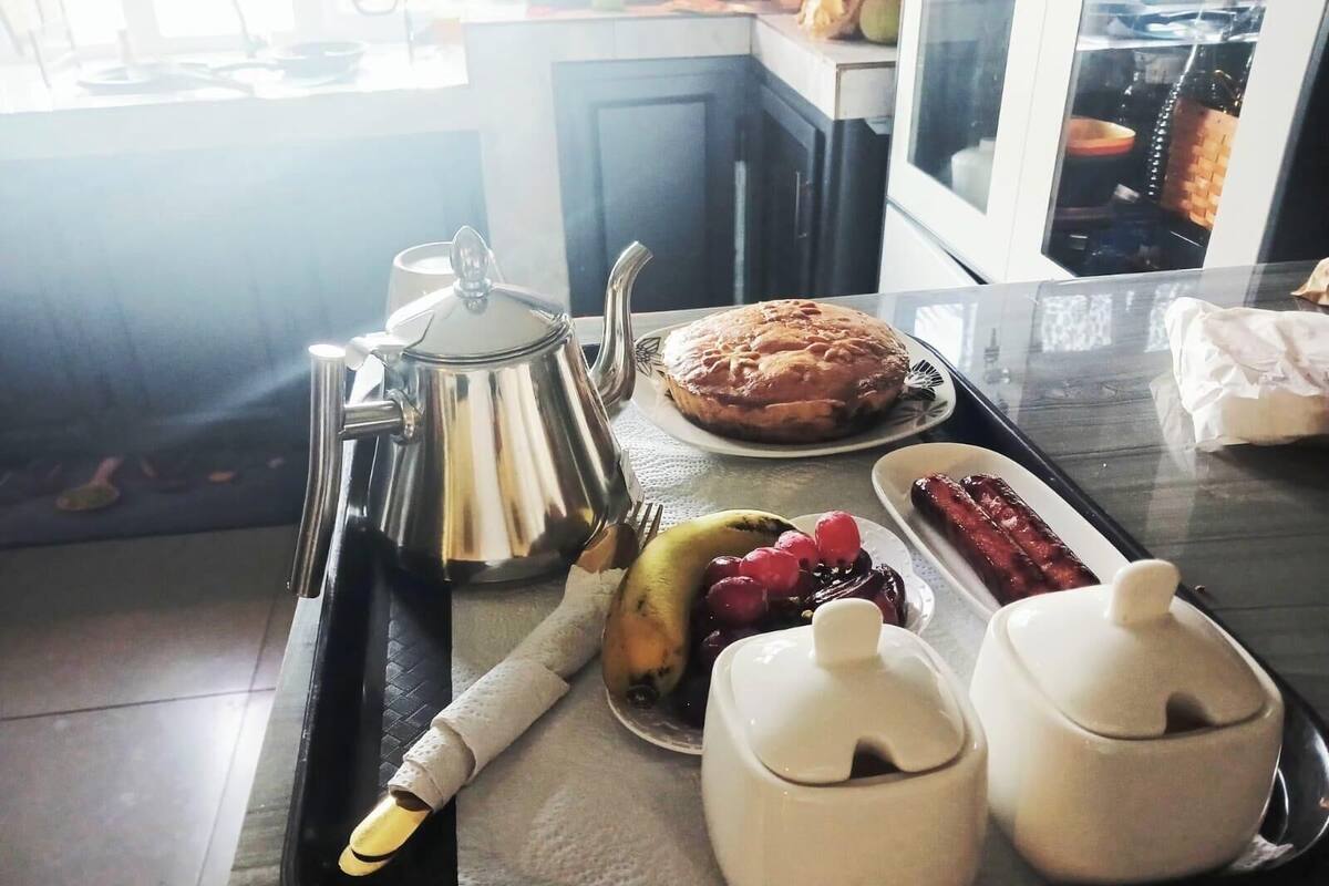 Un plateau de petit-déjeuner avec une théière, une miche de pain, une assiette de fruits et deux petits récipients.