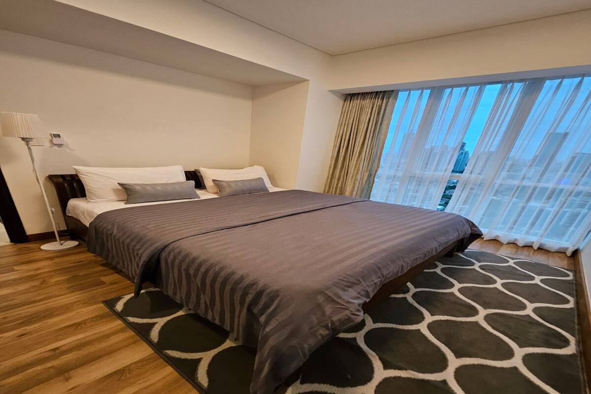 A hotel room with a large bed, a standing lamp, and a view of the city through sheer curtains.