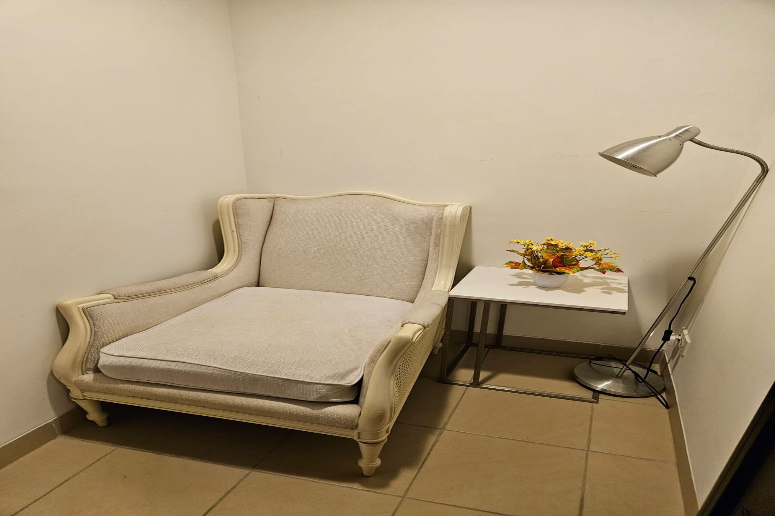Ein Eckzimmer mit weißem Ohrensessel, einem kleinen weißen Tisch mit Blumenvase und einer Stehlampe.