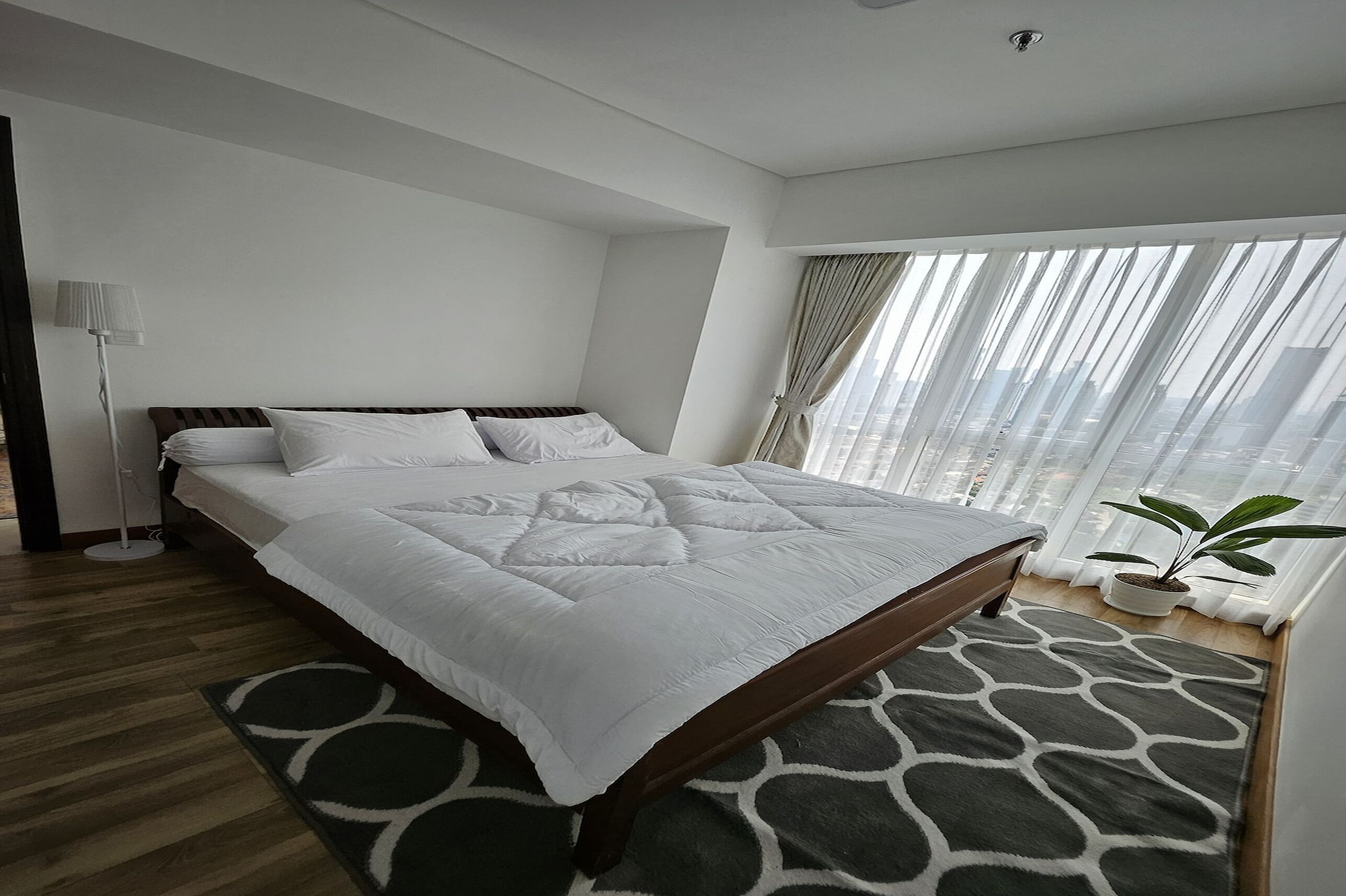 A modern hotel room with a large bed, a floor lamp, a potted plant, and a view of the city through sheer curtains.