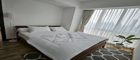 A modern hotel room with a large bed, a floor lamp, a potted plant, and a view of the city through sheer curtains.