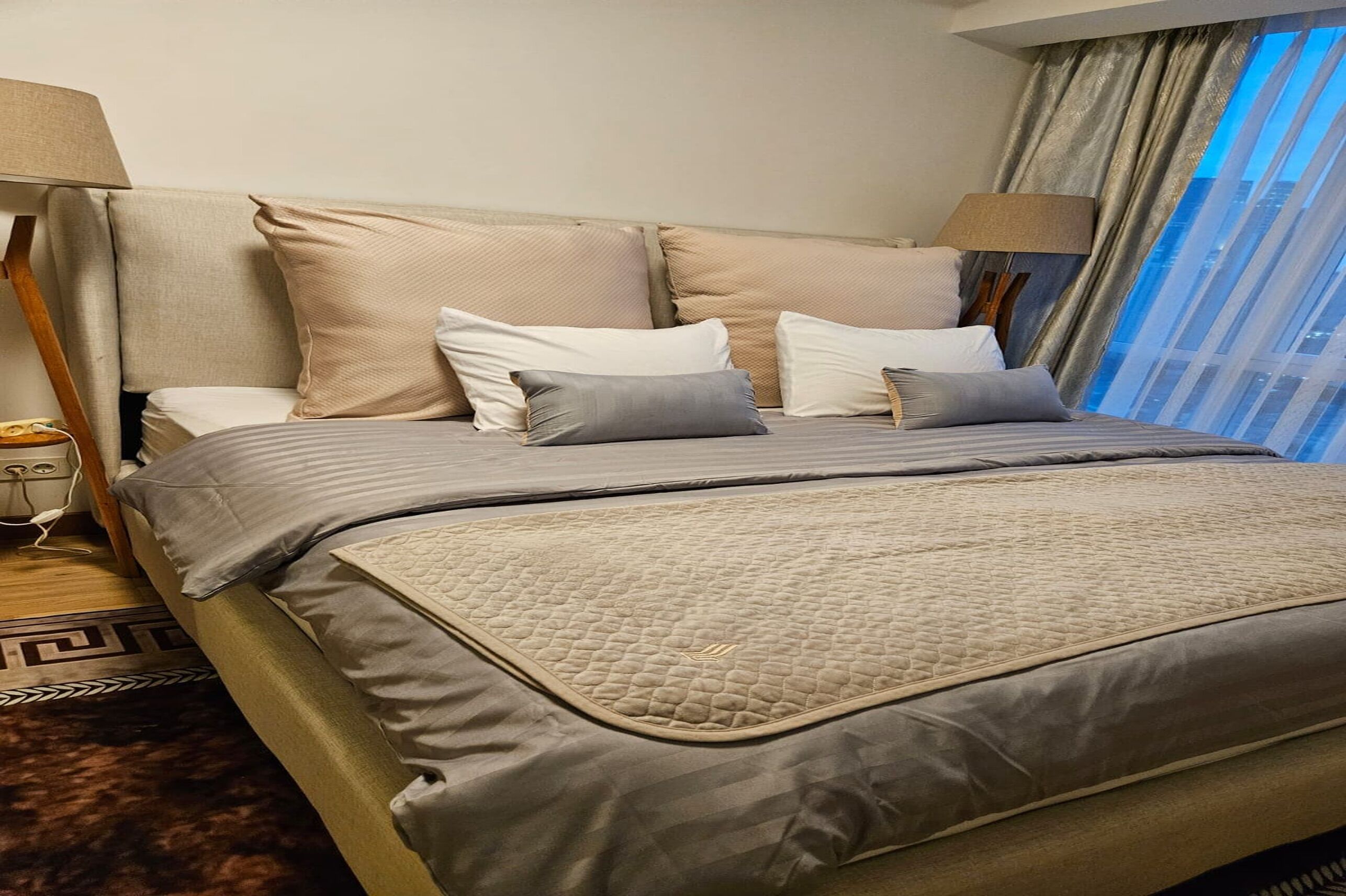 Habitación de hotel con una cama grande, cabecera y vistas de la ciudad a través de cortinas translúcidas.