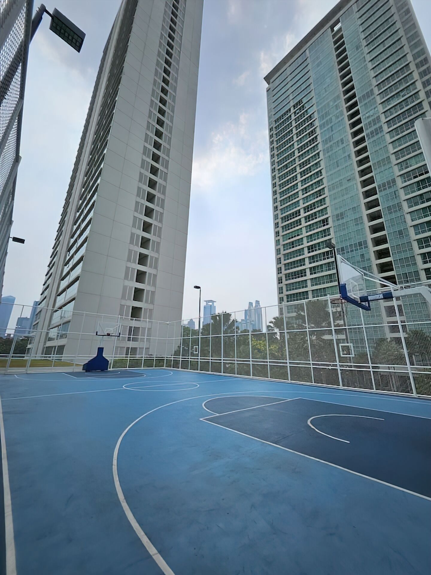 A basketball court with a blue surface and white lines, surrounded by tall buildings with reflective glass windows.