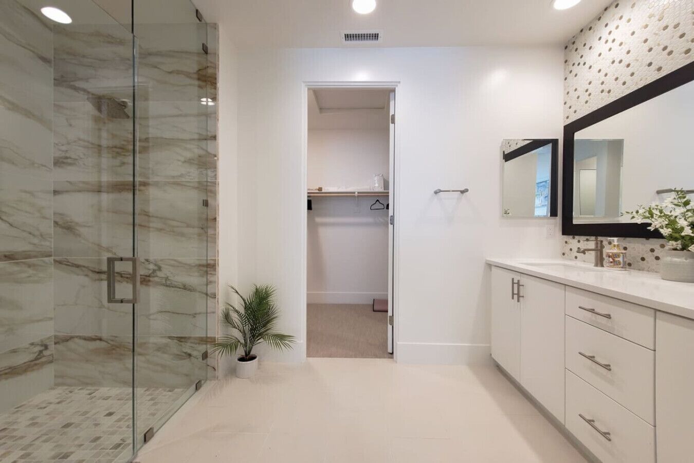 A modern bathroom with a glass shower enclosure, a walk-in closet, and a vanity with a large mirror.